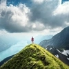 A person stands on a green mountain peak overlooking a lake and surrounding mountains under a cloudy sky.