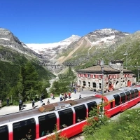 A red train travels through a mountain landscape with snow-covered mountains and a historic station building.