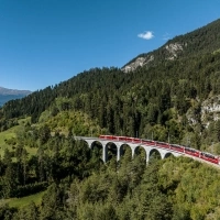 A red train crosses a viaduct in a mountainous and forested landscape under a clear blue sky.