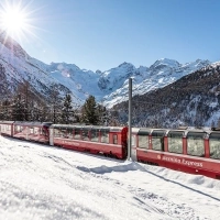 A red Bernina Express train travels through a snowy mountain landscape in sunshine.