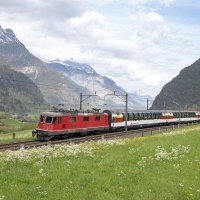 A red locomotive pulls a train through a green mountain landscape.