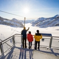 Eine Gruppe von drei Personen auf einer Aussichtsplattform mit Blick auf eine verschneite Berglandschaft.