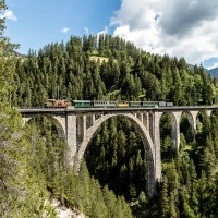 A historic train crosses a stone viaduct in the middle of a wooded valley.