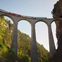 A red panoramic train crosses the impressive Landwasser Viaduct, an iconic structure nestled in a lush green mountain landscape.