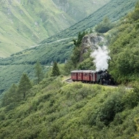 A steaming train travels through a green mountain landscape.