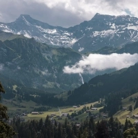 Un paesaggio montano con valli verdi, case sparse e cime innevate; nuvole e alberi in primo piano.