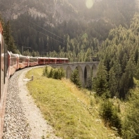 Un treno rosso attraversa un viadotto in un paesaggio montano boscoso.