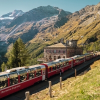 Ein roter Panoramazug fährt durch eine alpine Landschaft mit hohen Bergen und einem Steinhaus.