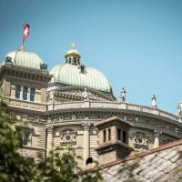 The image shows the Federal Palace in Bern with Swiss flags and a blue sky in the background.