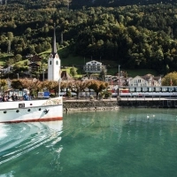Un ferry sur un lac, avec une église et des maisons dans un paysage de montagne verdoyant en arrière-plan.