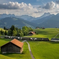 A landscape with green meadows, a moving train, and tall mountains in the background.