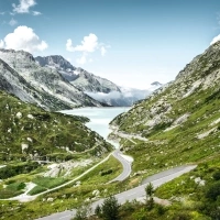 A winding mountain road through a green, rocky landscape with a lake and mountains in the background.