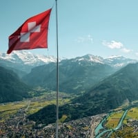 A group of people on a viewing platform overlooking a valley and mountains, next to a Swiss flag.