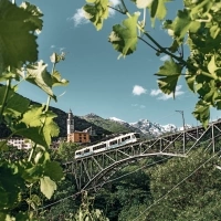 Un train traverse un pont entouré de feuilles vertes, avec un clocher d'église et des montagnes enneigées en arrière-plan.