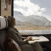 A person sitting in a train, looking out the window at a mountainous landscape with clouds and a lake.