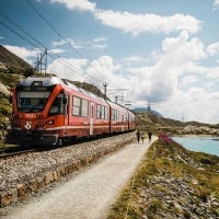 A red train travels along a turquoise lake and through an alpine landscape.