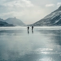 Two people ice skating on a frozen lake in front of a mountain landscape.