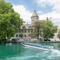 A long, narrow ferry on a river in front of a historic building and large trees.