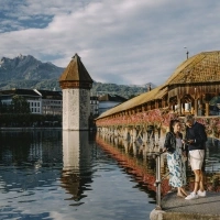 A pretty bridge with flowers, two people by the water, and mountains in the background.