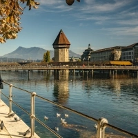 A woman walks along the shore, with the Chapel Bridge and the Water Tower in Lucerne in the background; mountains in the distance.