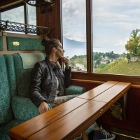 Eine Frau sitzt in einem Zugabteil und blickt nachdenklich aus dem Fenster auf eine Landschaft mit Weinbergen und einem Schloss.