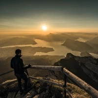 A person stands on a mountain, overlooking a lake and surrounding landscape at sunrise.