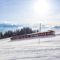 A red train travels through a snowy winter landscape with mountains in the background.