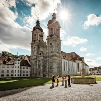 Eine barocke Kathedrale mit zwei Türmen, umgeben von alten Gebäuden und einem Platz mit Brunnen; Menschen spazieren bei sonnigem Wetter.