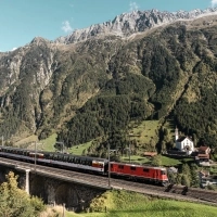 A red locomotive on a viaduct in front of a mountain landscape.