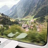 Vista da un finestrino del treno su un paesaggio alpino con un villaggio e montagne sullo sfondo.