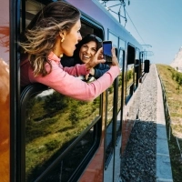 Two people leaning out of a train window taking a photo of a mountain landscape.