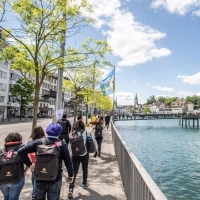 A group of people walking along a river in a city with historic buildings and a blue sky.