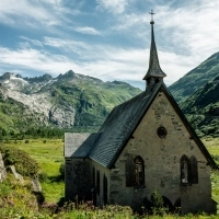 Una piccola cappella in pietra antica con un tetto a punta in un paesaggio alpino, circondata da colline verdi e montagne rocciose.