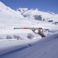 Ein roter Zug fährt durch eine tief verschneite Winterlandschaft, vorbei an schneebedeckten Bergen unter einem klaren, blauen Himmel.