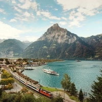 Paesaggio montano con lago, treno e nave in primo piano.