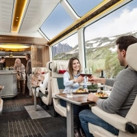 Interior view of a luxurious panoramic train with large windows; passengers enjoy an elegant dinner with views of the alpine landscape.