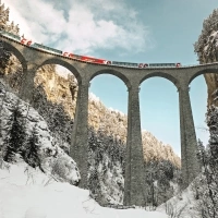 A red train travels over an impressive stone viaduct in the snowy mountains.