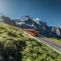 A red train travels through a green mountainous landscape with snow-covered peaks in the background.