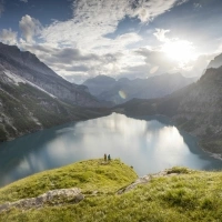 Un'ampia paesaggio montano con un lago tranquillo, circondato da alte scogliere e pendii boscosi; due persone stanno in un prato in primo piano.