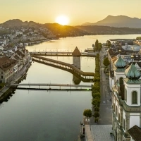 Sunset over a city with a bridge and river, mountains in the background.