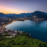 View of a lakeside city at dusk with lit streets and wooded hills in the foreground.