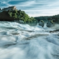 Wasserfall mit Schloss im Hintergrund, umgeben von üppiger Vegetation.