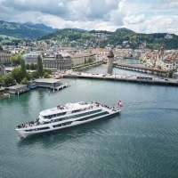 Un bateau avec le drapeau suisse navigue sur une rivière dans une ville à l'architecture médiévale avec un pont en bois, entourée de collines verdoyantes.