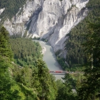 Blick auf eine Schlucht mit Fluss, bewaldete Hänge und Zug auf einer Brücke im Vordergrund.