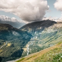 A winding mountain road snakes through a green valley under dramatic clouds.