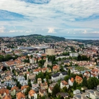 Aerial view of a cityscape with houses, trees, and a hill in the background under a partly cloudy sky.