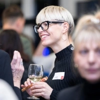 A smiling person with glasses holding a glass of white wine.