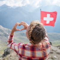 Eine Frau formt ein Herz mit ihren Händen vor einer Schweizer Flagge und Bergkulisse.
