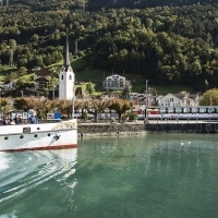 Un ferry sur un lac, avec une église et des maisons dans un paysage de montagne verdoyant en arrière-plan.