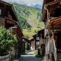Ein Dorfkern mit Holzchalets, auf deren Balkonen rote Blumen blühen, und im Hintergrund ein Berg.